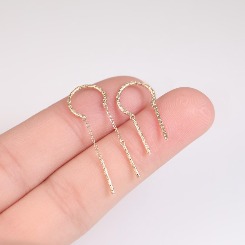 May include: A pair of delicate gold-colored earrings. Each earring features a textured, curved top connected to a chain and a textured bar. The earrings are displayed against a light background.