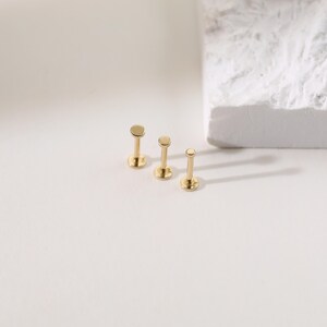 K&ouml;nnte beinhalten: Drei goldfarbene Lippen- oder Ohrstecker in verschiedenen Gr&ouml;&szlig;en. Jeder Stecker hat eine flache, kreisf&ouml;rmige Oberseite und einen Stift zum Einsetzen. Die Stecker werden auf einer wei&szlig;en Oberfl&auml;che mit einem strukturierten wei&szlig;en Block im Hintergrund pr&auml;sentiert.