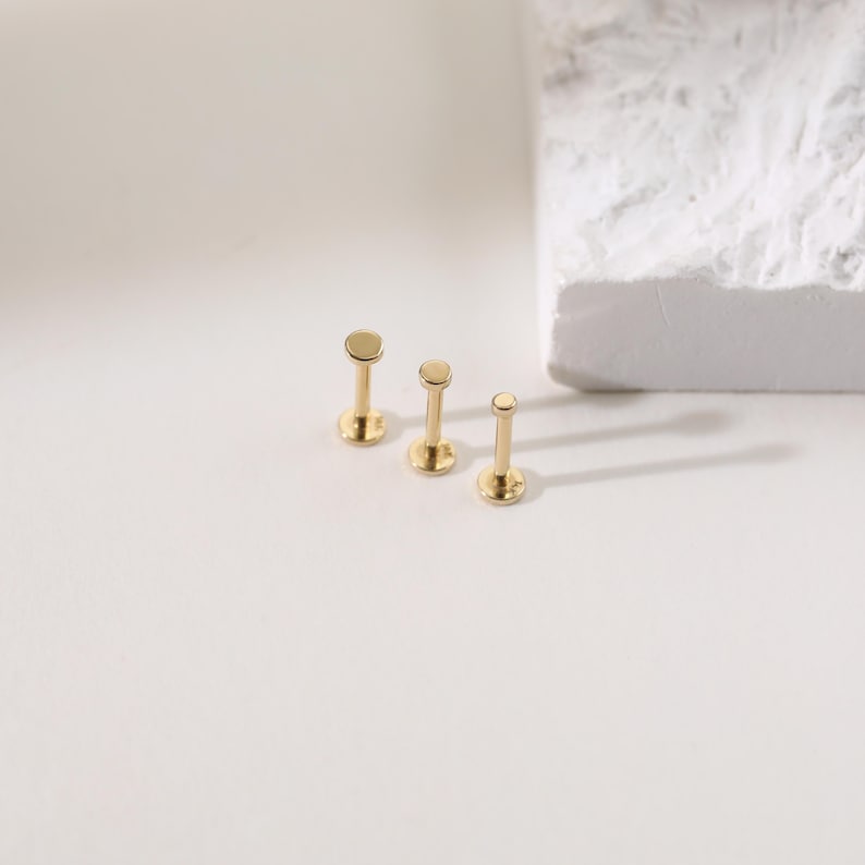 K&ouml;nnte beinhalten: Drei goldfarbene Lippen- oder Ohrstecker in verschiedenen Gr&ouml;&szlig;en. Jeder Stecker hat eine flache, kreisf&ouml;rmige Oberseite und einen Stift zum Einsetzen. Die Stecker werden auf einer wei&szlig;en Oberfl&auml;che mit einem strukturierten wei&szlig;en Block im Hintergrund pr&auml;sentiert.