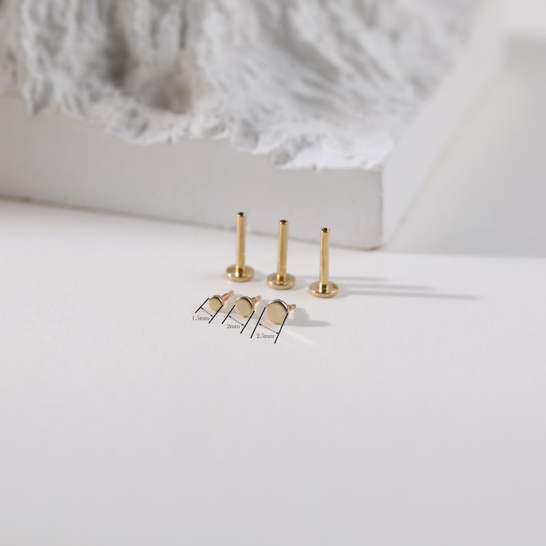 K&ouml;nnte beinhalten: Goldfarbene K&ouml;rperschmuck-Ohrstecker in verschiedenen Gr&ouml;&szlig;en. Das Bild zeigt drei Ohrstecker mit flachen R&uuml;ckseiten und drei mit gebogenen R&uuml;ckseiten, mit den Ma&szlig;en 1,5 mm, 2 mm und 2,5 mm. Der Schmuck wird auf einer wei&szlig;en Oberfl&auml;che pr&auml;sentiert.