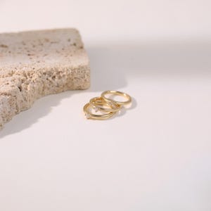 May include: Three gold rings with small, clear gemstones are displayed on a white surface. One ring has a single gemstone, while another has multiple. The rings are positioned near a textured, beige stone.