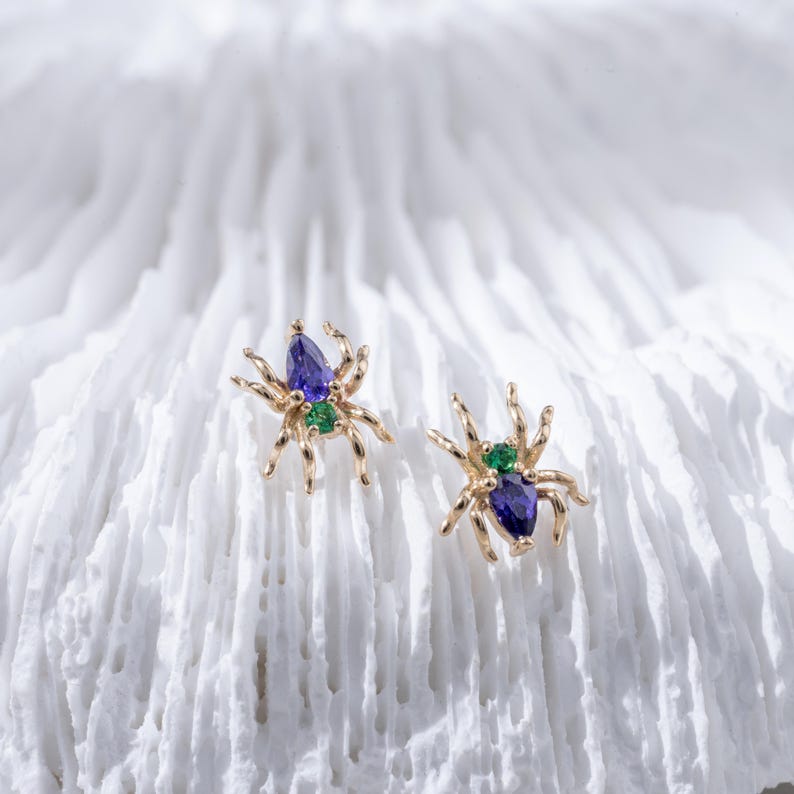 May include: A pair of gold spider earrings with blue and green gemstones. The earrings are on a white coral-like surface.