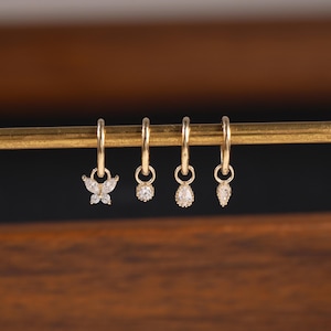 May include: Four gold hoop earrings with dangling charms. One earring features a butterfly design, while the others have round and teardrop-shaped clear gemstones. The earrings are displayed on a gold bar against a dark background.