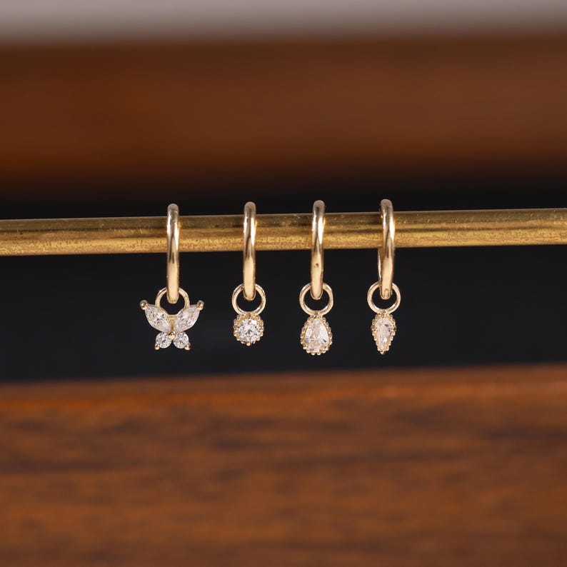 May include: Four gold hoop earrings with dangling charms. One earring features a butterfly design, while the others have round and teardrop-shaped clear gemstones. The earrings are displayed on a gold bar against a dark background.
