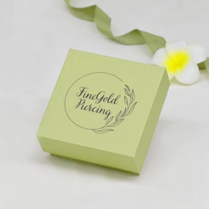 May include: A light green square box with a circular logo that says "FineGold Piercing" in a black cursive font. The logo is surrounded by a wreath of leaves.