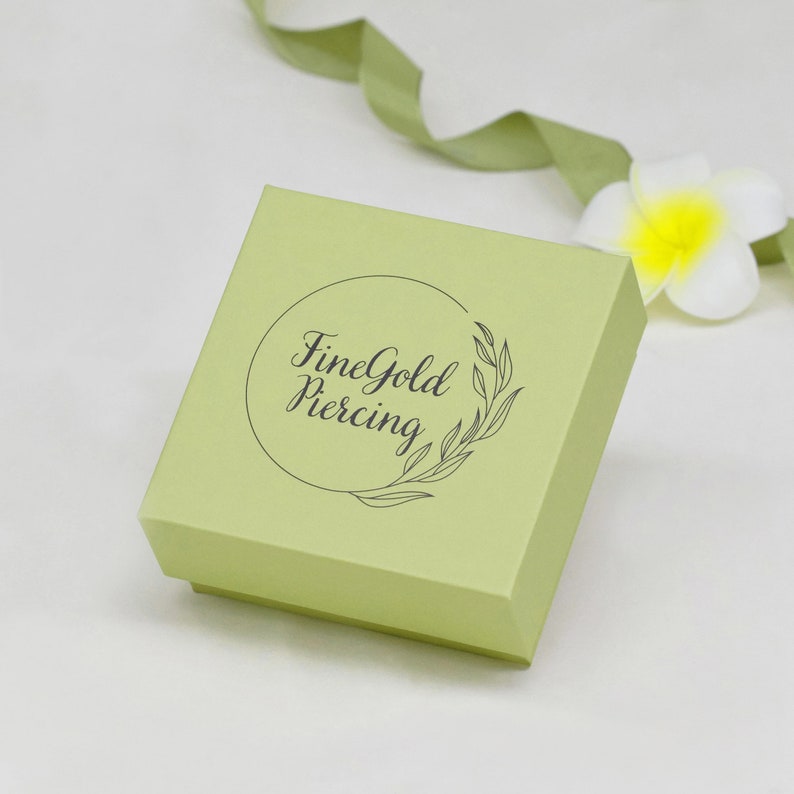 May include: A light green square box with a circular logo that says "FineGold Piercing" in a black cursive font. The logo is surrounded by a wreath of leaves.