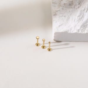 K&ouml;nnte beinhalten: Drei goldfarbene Ohrstecker mit flachen Oberteilen in verschiedenen Gr&ouml;&szlig;en sind auf einer wei&szlig;en Oberfl&auml;che angeordnet. Jeder Ohrring hat einen Stecker und eine kreisf&ouml;rmige Oberseite. Im Hintergrund befindet sich ein strukturierter wei&szlig;er Block mit weichen Schatten.