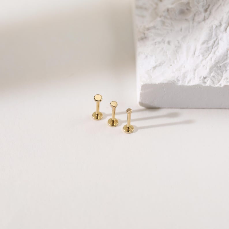 K&ouml;nnte beinhalten: Drei goldfarbene Ohrstecker mit flachen Oberteilen in verschiedenen Gr&ouml;&szlig;en sind auf einer wei&szlig;en Oberfl&auml;che angeordnet. Jeder Ohrring hat einen Stecker und eine kreisf&ouml;rmige Oberseite. Im Hintergrund befindet sich ein strukturierter wei&szlig;er Block mit weichen Schatten.