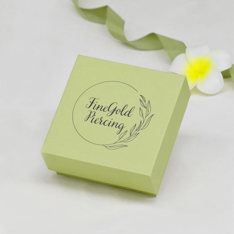 K&ouml;nnte beinhalten: Hellgr&uuml;ne quadratische Schachtel mit dem Text "FineGold Piercing" in einem kreisf&ouml;rmigen Design. Die Schachtel ist mit einem Bl&auml;tterzweig-Design verziert. Eine wei&szlig;e Blume mit gelbem Zentrum und ein gr&uuml;nes Band befinden sich im Hintergrund.