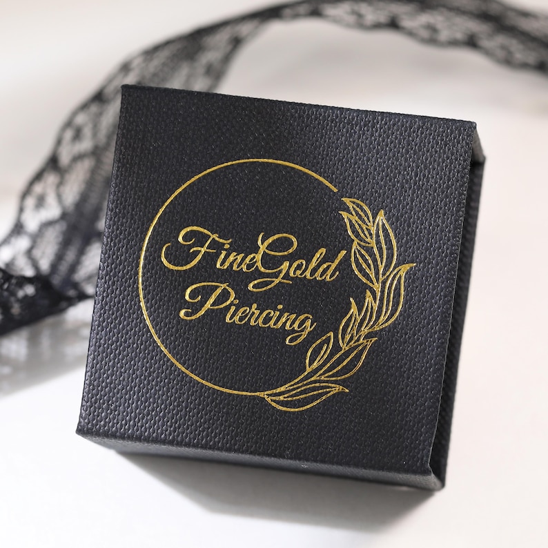 May include: A square black jewelry box with a textured surface. The top of the box features a gold foil logo with the text FineGold Piercing enclosed in a circular frame with a decorative leaf design on the right side of the circle.