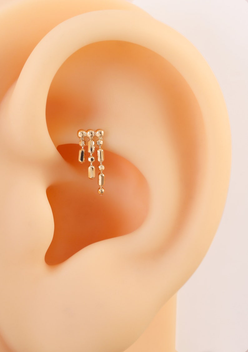 May include: Gold ear cuff with a dangling chain of small beads and bars. The cuff is worn in the upper cartilage of the ear.