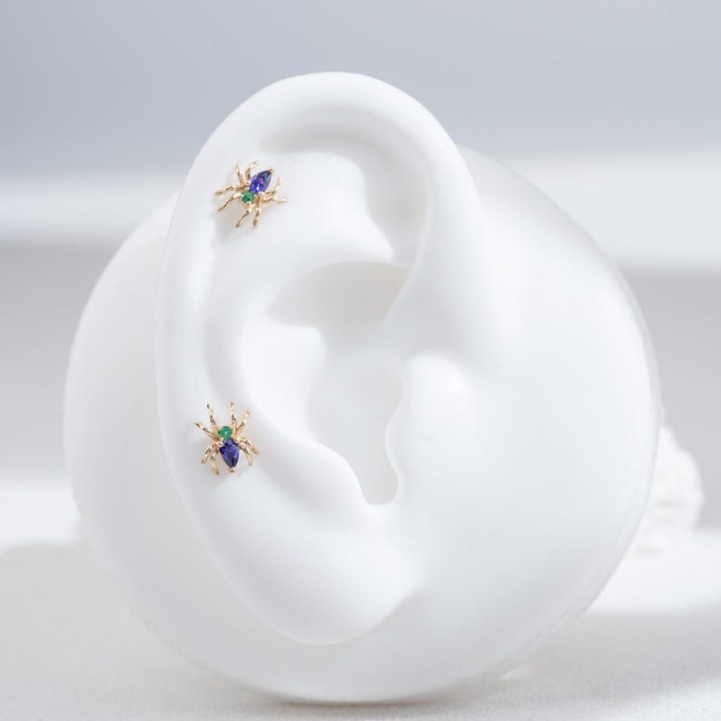 May include: A pair of gold spider earrings with green and blue gemstones. The earrings are shown on a white mannequin ear.