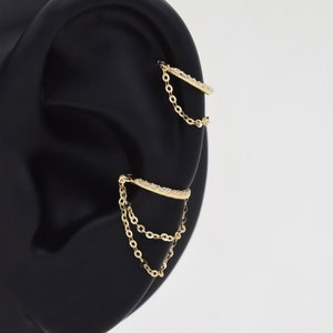 May include: Gold ear cuff with a chain dangling from it. The cuff is decorated with small clear stones.