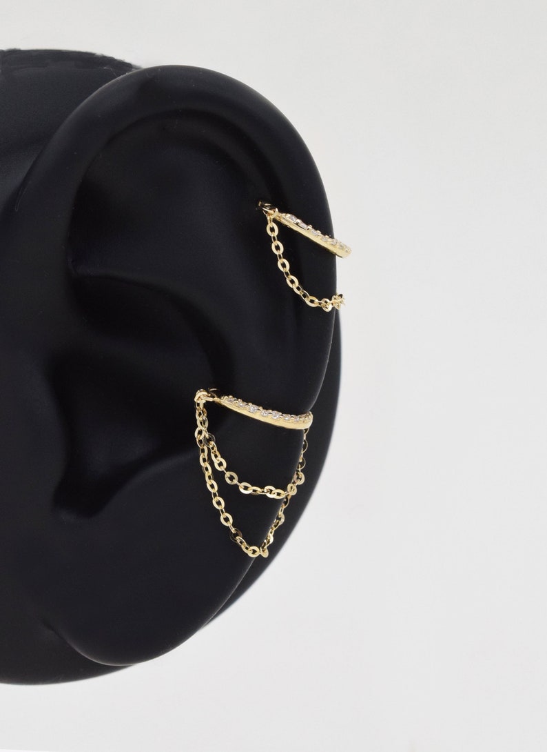 May include: Gold ear cuff with a chain dangling from it. The cuff is decorated with small clear stones.