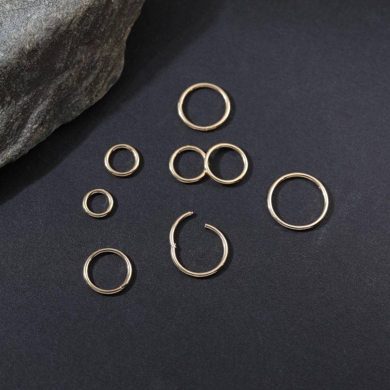 May include: A collection of seven gold-toned circular rings of varying sizes arranged on a dark surface. The rings are thin, smooth, and metallic, with one ring featuring a visible hinge mechanism for opening and closing the circular band.