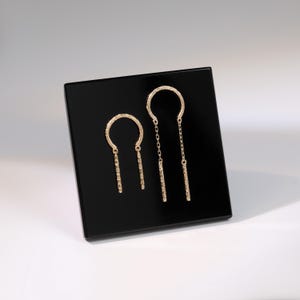 May include: A pair of gold-colored earrings displayed on a black square. The earrings feature a textured half-circle design with a dangling bar. One earring has a chain. The earrings are presented on a black surface against a white background.