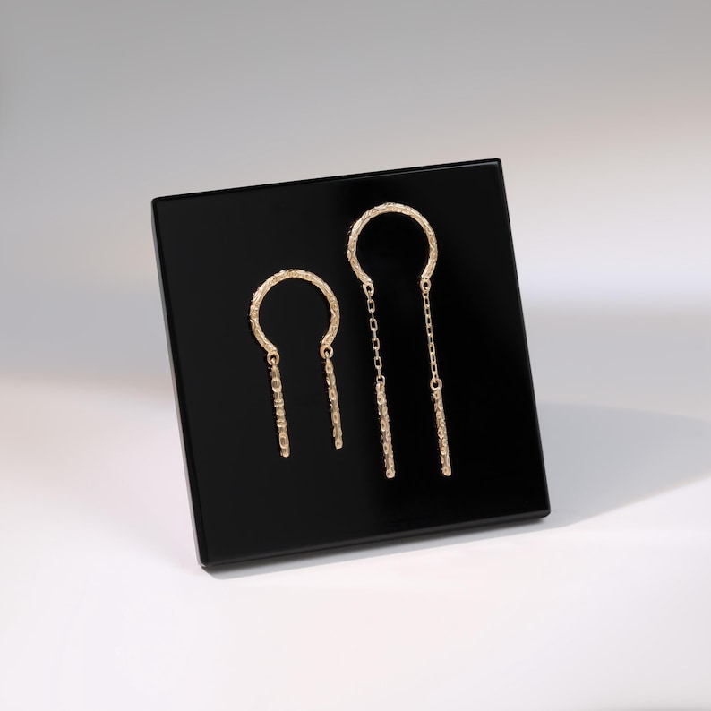 May include: A pair of gold-colored earrings displayed on a black square. The earrings feature a textured half-circle design with a dangling bar. One earring has a chain. The earrings are presented on a black surface against a white background.