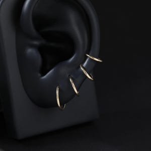 May include: A set of four thin, polished gold hoop earrings arranged in a vertical row along the outer edge of a black ear-shaped display model. Each hoop is circular, smooth, and features a simple, continuous design with a reflective metallic finish.