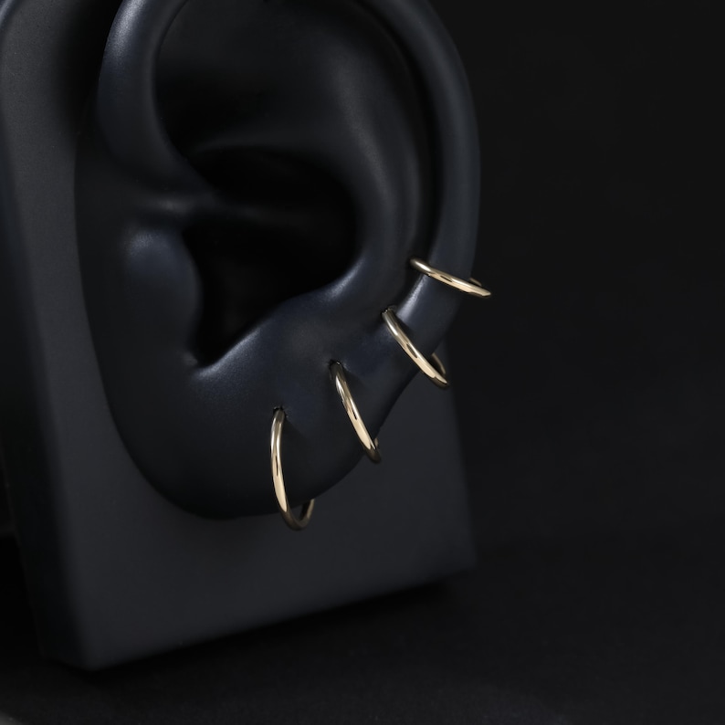 May include: A set of four thin, polished gold hoop earrings arranged in a vertical row along the outer edge of a black ear-shaped display model. Each hoop is circular, smooth, and features a simple, continuous design with a reflective metallic finish.