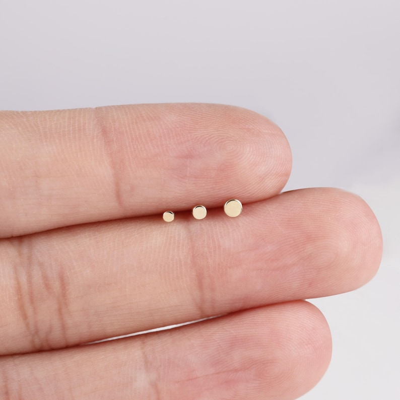 K&ouml;nnte beinhalten: Drei kleine, goldfarbene Ohrstecker in verschiedenen Gr&ouml;&szlig;en werden auf dem Finger einer Person gezeigt. Die Ohrringe sind einfache, runde Ohrstecker. Der kleinste Ohrring hat einen Durchmesser von ca. 0,25 cm, der gr&ouml;&szlig;te von ca. 0,5 cm.