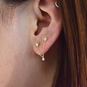 May include: A pair of gold star-shaped earrings with small clear stones. The earrings are connected by a gold chain with a small clear stone dangling from the end.