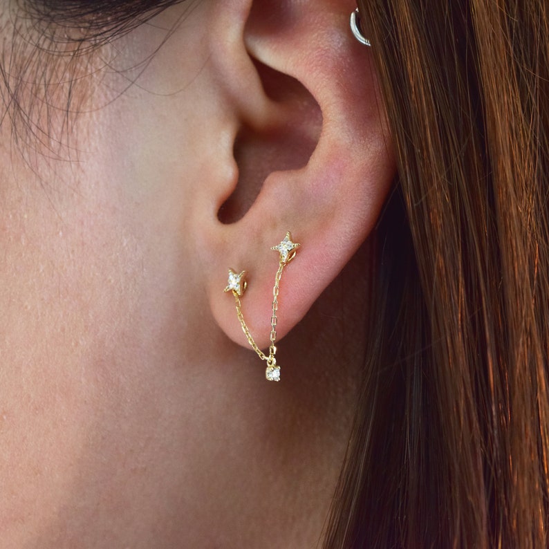 May include: A pair of gold star-shaped earrings with small clear stones. The earrings are connected by a gold chain with a small clear stone dangling from the end.