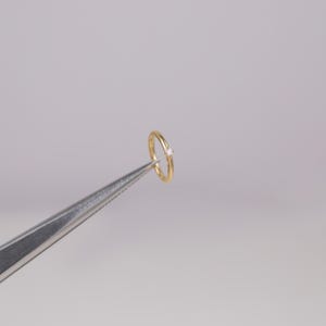 May include: A delicate gold ring with a small, clear gemstone. The ring is held by a pair of silver tweezers against a plain, light gray background. The ring's band is thin and polished.