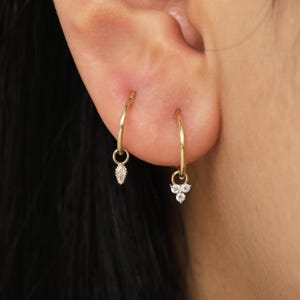 May include: A pair of gold hoop earrings. One earring features a dangling marquise-cut gemstone, while the other has three round gemstones. The earrings are worn on a person's ear.