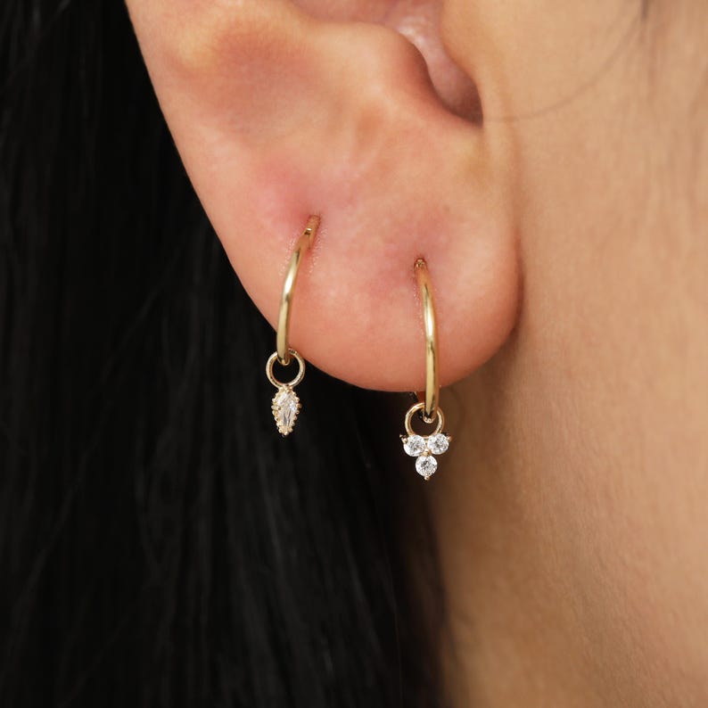 May include: A pair of gold hoop earrings. One earring features a dangling marquise-cut gemstone, while the other has three round gemstones. The earrings are worn on a person's ear.