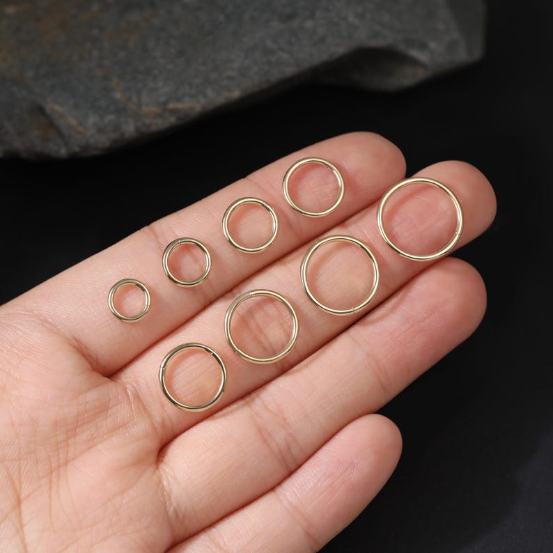 May include: A collection of eight smooth, circular gold-toned metal rings arranged in a graduated size sequence. The rings are thin, polished, and feature a simple, continuous band design, displayed in a variety of diameters from small to large.