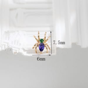 May include: Gold spider charm with a purple and green gemstone. The charm is 6mm wide and 7.5mm tall.