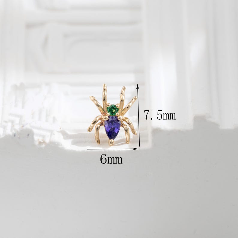 May include: Gold spider charm with a purple and green gemstone. The charm is 6mm wide and 7.5mm tall.