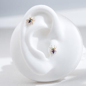 May include: A pair of gold spider earrings with green and blue gemstones. The earrings are shown on a white mannequin ear.