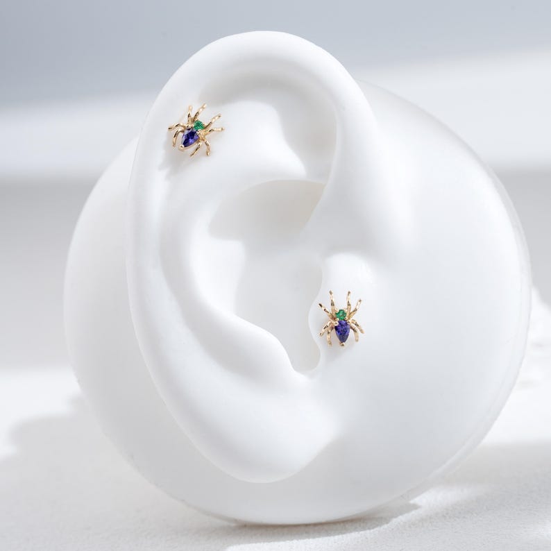 May include: A pair of gold spider earrings with green and blue gemstones. The earrings are shown on a white mannequin ear.