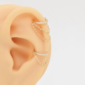 May include: Gold ear cuff with a chain dangling from it. The cuff is decorated with small clear stones.