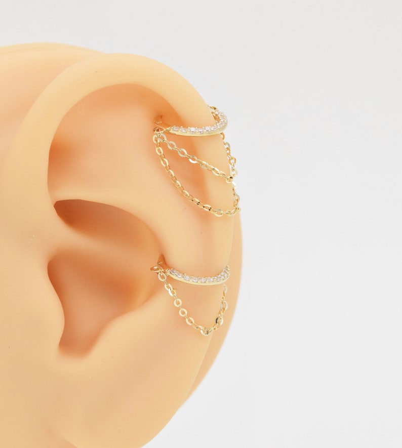 May include: Gold ear cuff with a chain dangling from it. The cuff is decorated with small clear stones.