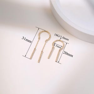 May include: Two gold-colored earrings with a unique design. One earring is 31mm long, the other is 20mm long. The earrings have a circular top with a dangling bar. The smaller earring's circle is 8.5mm in diameter. The gauge is 18G/1.0mm.