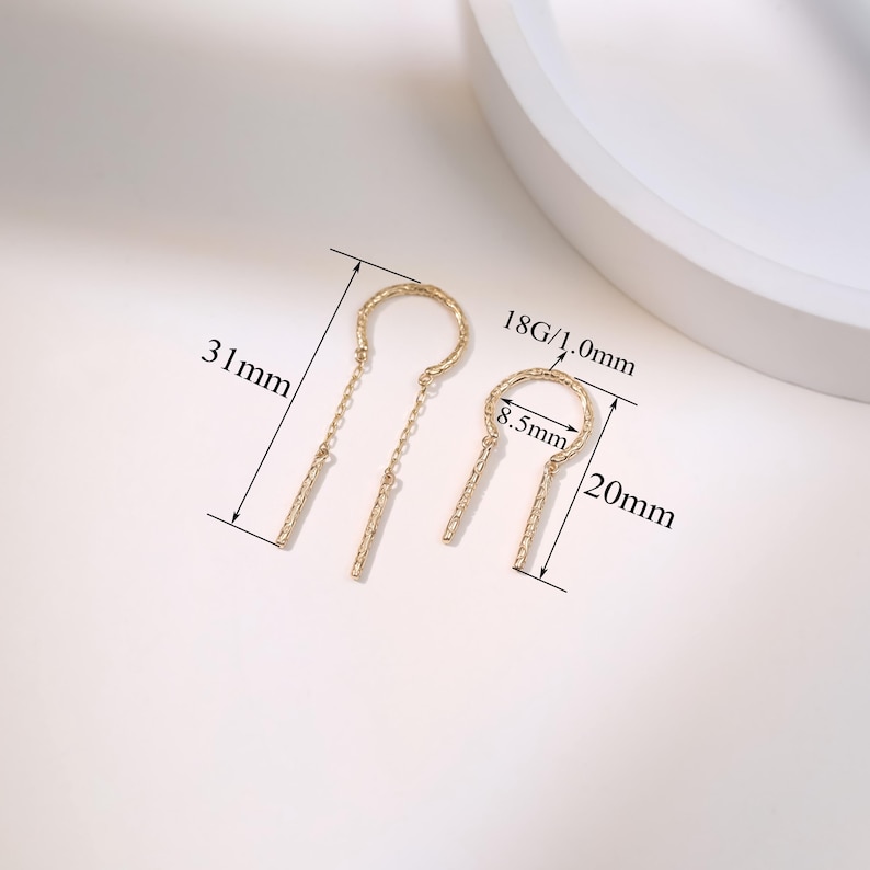 May include: Two gold-colored earrings with a unique design. One earring is 31mm long, the other is 20mm long. The earrings have a circular top with a dangling bar. The smaller earring's circle is 8.5mm in diameter. The gauge is 18G/1.0mm.