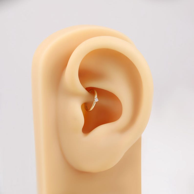 May include: A gold-colored hoop earring with a small, clear gemstone. The earring is set in a model ear, showcasing the jewelry's design and placement. The earring is a simple, elegant piece.