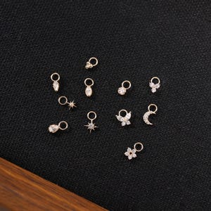 May include: A collection of small, gold-colored charms with various designs, including teardrops, stars, flowers, butterflies, and moons. Each charm has a small loop for attaching to jewelry. The charms are displayed on a black surface.