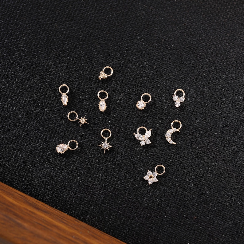 May include: A collection of small, gold-colored charms with various designs, including teardrops, stars, flowers, butterflies, and moons. Each charm has a small loop for attaching to jewelry. The charms are displayed on a black surface.