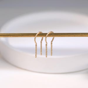 May include: A pair of gold-colored drop earrings. Each earring features a textured, curved top and a straight, textured bar that hangs down. The earrings are displayed on a gold-colored bar against a soft, white background.