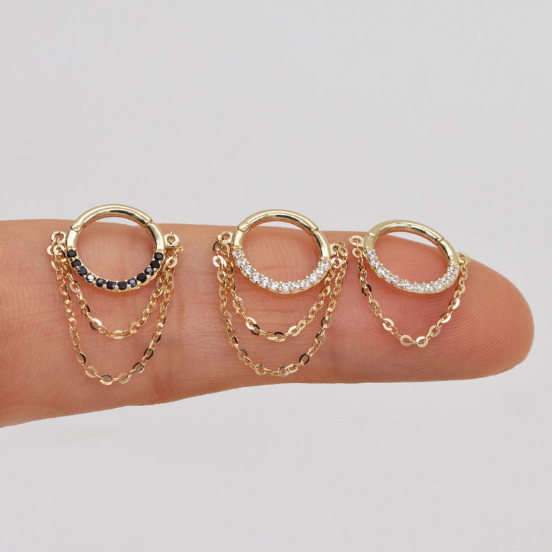 May include: Three gold hoop earrings with dangling chains. The hoops are set with small black or white stones. The chains are layered and have a delicate, feminine look.