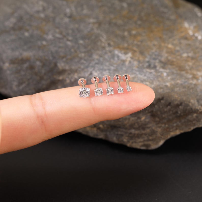 Pu&ograve; includere: Cinque orecchini a perno d'argento con pietre di zircone cubico trasparenti. Gli orecchini sono disposti in fila su un dito.