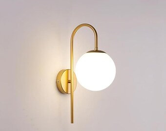 May include: A wall-mounted light fixture featuring a circular brass-toned metal base and a slender, curved arm that extends upward and downward. A spherical, opaque white glass globe shade is attached to the arm, emitting a soft, warm glow.