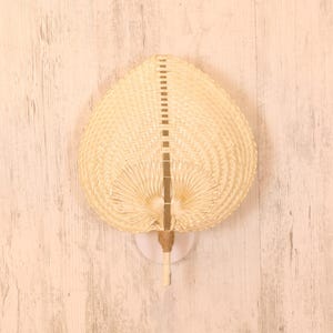 May include: A hand fan made of woven natural fibers, featuring a heart-shaped design. The fan has a light brown color and a handle wrapped with twine. The fan is mounted on a white base.