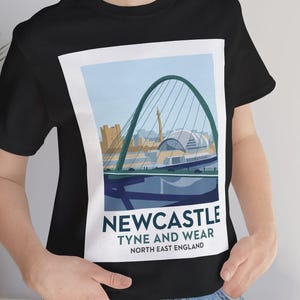 Puede incluir: Camiseta negra con un gráfico del puente Tyne en Newcastle, Inglaterra, dentro de un marco blanco. El gráfico incluye el texto "Newcastle Tyne and Wear North East England". El puente es verde, sobre un cielo azul claro.