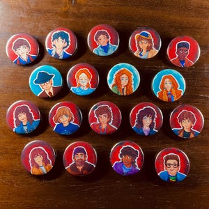 May include: A collection of twenty round pinback buttons featuring colorful illustrations of characters. Each button has a red border and a unique character portrait, with varying skin tones, hair colors, and clothing styles. The buttons are arranged on a brown surface.