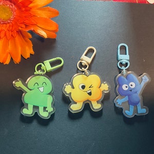 May include: Three colorful cartoon character keychains with different expressions. The characters are green, yellow, and blue. Each keychain has a silver or blue metal ring.
