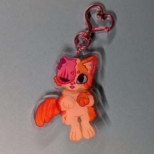 May include: An acrylic keyring featuring a cartoon orange tabby cat with a pink brain-like patch on its head, one large dark eye, and a heart-shaped pink metal clasp. The cat has orange stripes on its tail and ears, with a light cream-coloured body.